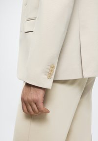 Bežinė švarkas su lygaus paviršiaus, su keturiais smėlio spalvos mygtukais ant rankovių ir notch apykakle. Medžiaga atrodo lengva ir pritaikyta.