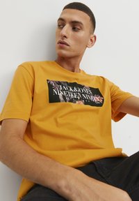 T-shirt di cotone giallo con un motivo floreale nero e testo bianco. Modello che indossa pantaloni neri, seduto su uno sfondo bianco.