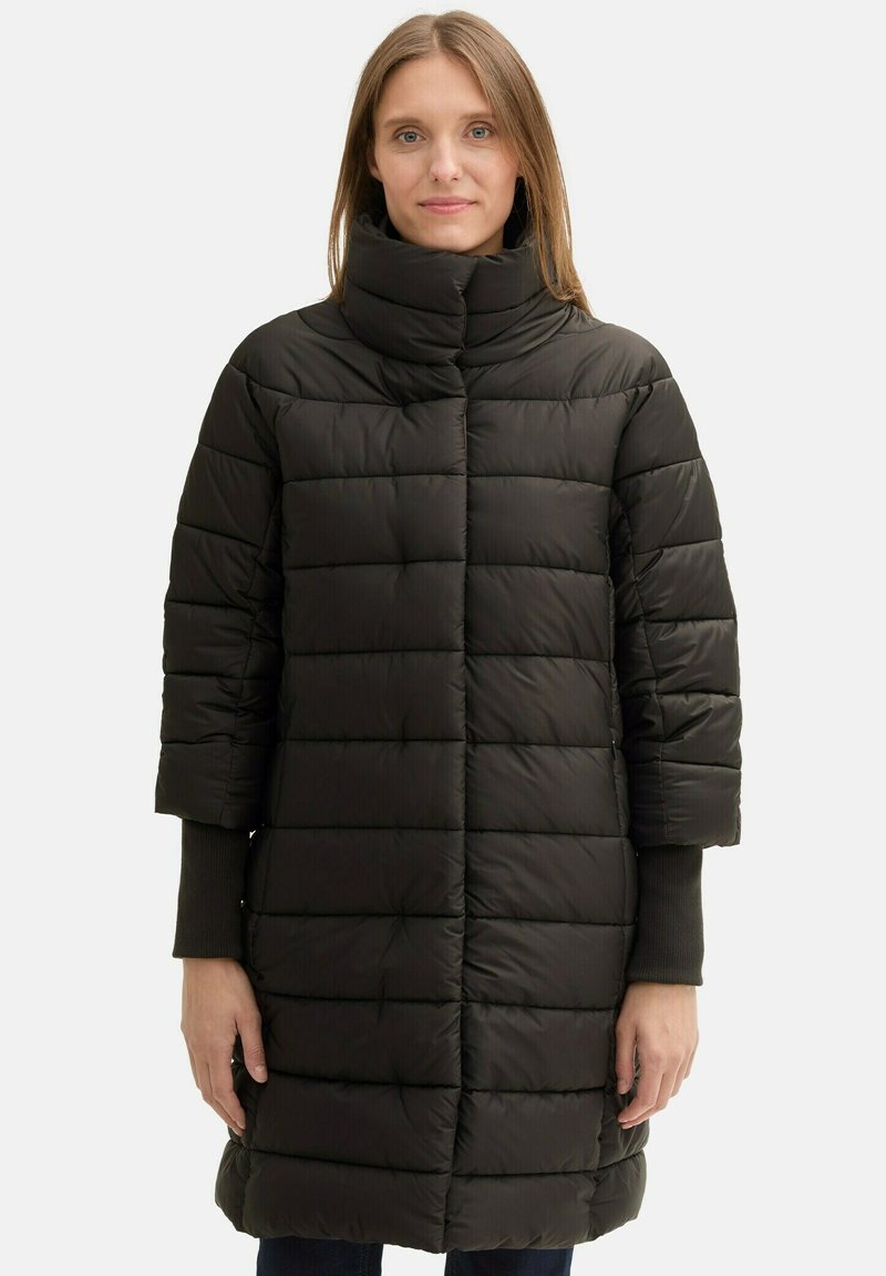 Crna, quilted puffer jakna s visokim ovratnikom, kratkim rukavima i ravnim krojem koji pada do sredine bedara. Glatka tekstura, minimalne metalne detalje.