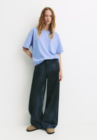 Tricou supradimensionat de culoare albastru deschis, fabricat din bumbac, asortat cu blugi negri cu croi larg. Modelul poartă pantofi slip-on de culoare bej.