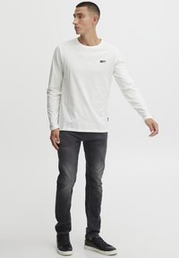 Hvid langærmet skjorte med rund halsudskæring, forsynet med et lille logo på brystet. Sat sammen med mørkegrå jeans og sorte sneakers.