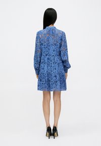 Μπλε δαντελένιο φόρεμα με λουλουδένιο μοτίβο, μακριά μανίκια και σGathered μέση, διαθέτει ψηλό γιακά και φούστα με επίπεδα. Φορεμένο με μαύρες γόβες.