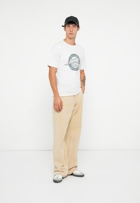 Tricou alb cu mânecă scurtă, cu imprimeu grafic verde, asortat cu pantaloni bej largi și o șapcă neagră. Încălțămintea include pantofi sport albi și gri.