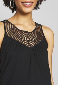 Černý bezrukávový top s texturovanou krajkovou částí s kruhovými vzory na výstřihu a volným, splývavým designem.