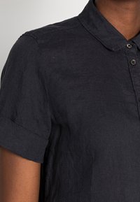 Camicia di lino blu scuro con colletto, maniche corte arrotolate e chiusura con bottoni. Il tessuto ha una finitura leggermente strutturata.