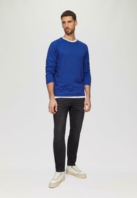 Blaues Langarm-Sweater mit Rundhalsausschnitt, über einem weißen Hemd getragen, kombiniert mit schwarzen Slim-Fit-Jeans und beigen Sneakers. Schlichte Texturen.