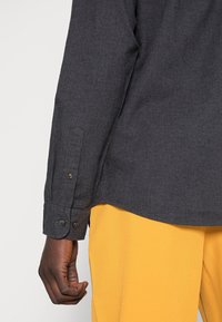 Tmavě šedý rukáv dlouhého trička s knoflíky přes hořčicové kalhoty na tmavé pokožce paže na jednobarevném pozadí.