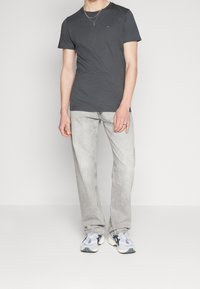 Graues Baumwoll-T-Shirt mit Rundhalsausschnitt, das ein kleines Logo auf der Brust trägt. Kombiniert mit hellgrauen Jeans und weißen Turnschuhen mit schwarzen Akzenten.