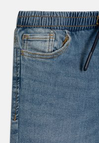 Svetlo modri jeans hlače z elastičnim pasom, šivanimi žepi in detajlom z vrvico. Imajo gladko teksturo in klasičen kroj.