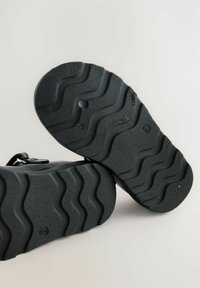 Semelles de chaussures noires avec un motif texturé et ondulé. Matériau en caoutchouc durable avec une forme ronde, présentant un petit trou près du centre.