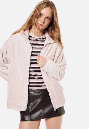 Jeune femme portant une veste peluche rose claire sur une chemise rayée et une jupe en cuir noire, tenant la fermeture éclair de la veste.