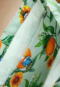 Portofel floral cu căptușeală din material verde, cu flori portocalii și galbene, frunze și un design de personaj de desene animate. Marginile cusute sunt vizibile.