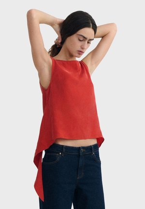 Woman adjusting hair wearing sleeveless red top with asymmetrical hem and dark blue jeans against plain background.