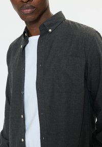 Hombre vistiendo una camisa de botones gris oscuro con botones blancos sobre una camiseta blanca lisa con cuello redondo, recortado a la altura de los hombros y la boca.