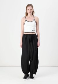 Fehér, fekete szegélyű, rövidített tank top, amelyet laza, fekete nadrággal és fekete sportcipővel párosítanak; tetoválás látható a karon.