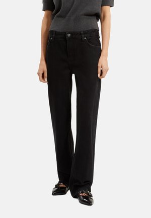 Pantalon en velours côtelé noir à jambes larges, avec une texture lisse, des poches avant et une fermeture à bouton. Accompagné de chaussures noires.