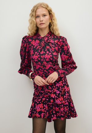 Kobieta o kręconych blond włosach, ubrana w czarną sukienkę w różowy kwiatowy wzór, z bufkami na rękawach i falbanką na dole, stojąca na jednolitym tle.