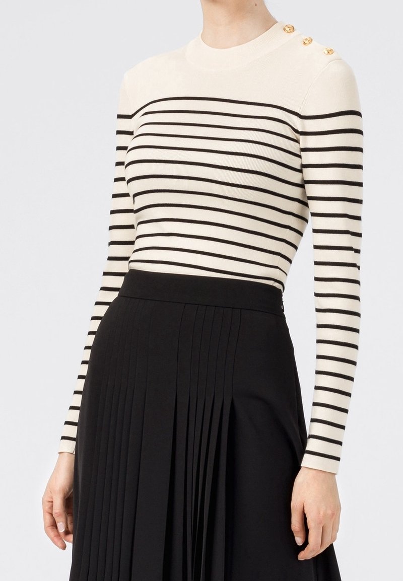 Femme portant un haut blanc à manches longues avec des rayures horizontales noires et des boutons dorés sur l'épaule, associé à une jupe plissée noire.