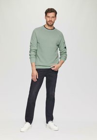 Lichtgroene gebreide trui met ribbelmanchetten, gecombineerd met donkere spijkerbroek en witte sneakers. De trui heeft een ronde hals en een zijzak.