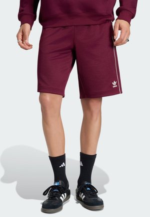 Mężczyzna w bordo spodenkach Adidas, czarnych skarpetkach Adidas i czarnych butach sportowych Adidas stojący na szarym tle.