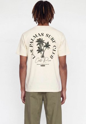 Homme avec des dreadlocks portant un t-shirt crème avec un motif de palmier noir et le texte Las Palmas Surf Club Costa Rica au dos.