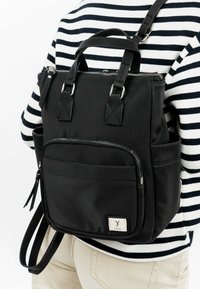 Black backpack with vertical ribbed texture, top handles, adjustable straps, front zipper pocket, and a small label on the side.