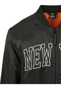 Chaqueta bomber negra con un interior naranja, con "NEW YORK" en texto blanco en el pecho. Fabricada con un tejido suave y brillante.