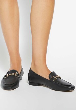 Paire de mocassins en cuir noir avec détail chaîne dorée portés pieds nus sur un fond blanc uni.