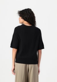 Maglione nero a maniche corte in un morbido tessuto a maglia con scollo a costine e orlo, indossato sopra pantaloni beige larghi.