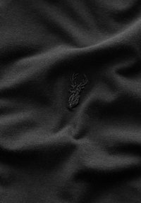 Textil negro con un discreto logo de ciervo bordado. La tela es suave, con un ligero brillo y un patrón de tejido ajustado.