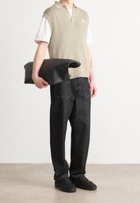 Ärmelloser Strickpullover in hellbeige über einem weißen Hemd mit Kragen, kombiniert mit dunklen Denim-Hosen und schwarzen Sneakers; hält eine schwarze Tasche.