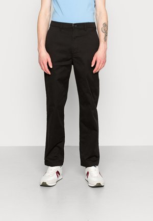 Schwarze Baumwollhosen mit geradem Schnitt, ausgestattet mit einem Bund, vorderen Taschen und einer glatten Textur. Kombiniert mit weißen Sneakern.