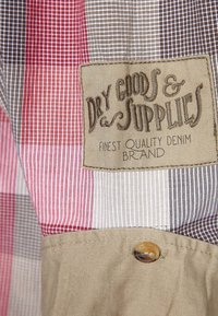 Kraagshirt van patchworkstof; heeft rode, witte en bruine strepen. Label leest "Dry Goods & Supplies" in gestikt bruine tekst.