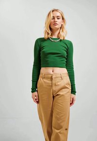 Top cropped verde com nervuras e mangas longas, combinado com calças beje de cintura alta. Um colar de corrente prateada complementa o conjunto.