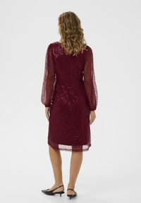 Rochie burgundă cu mâneci lungi din tul, model brodat complex, croială până la genunchi și talie ajustată, asortată cu pantofi slingback negri.