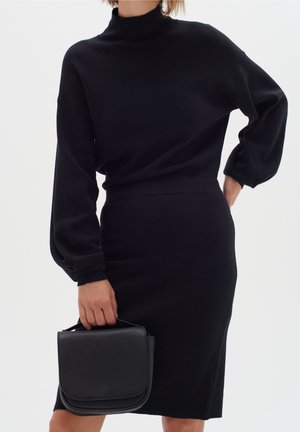 Frau trägt ein schwarzes Langarmkleid und hält eine kleine schwarze Handtasche vor einem schlichten weißen Hintergrund.