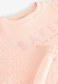 Sweat-shirt rose avec des détails en dentelle sur les côtés, col côtelé et lettrage en relief qui épelle "BAKER" dans une teinte plus claire.
