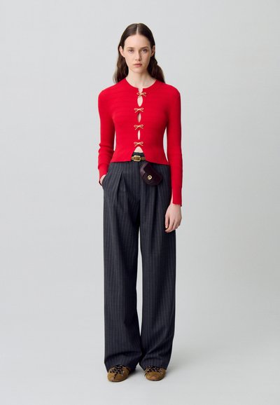 Top a maniche lunghe rosso a coste con bottoni a forma di fiocco in tono oro e dettaglio a taglio, abbinato a pantaloni larghi grigio a righe e scarpe marroni.
