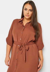 Braunes Hemdblusenkleid mit Bindegürtel, ausgestattet mit einem Kragen und hochgekrempelten Ärmeln. Glatte Textur und lockere Passform.