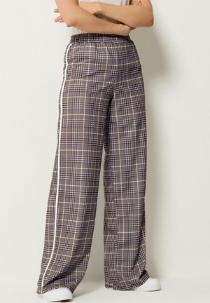 Person trägt karierte, hochtaillierte Hosen mit weitem Bein, weißen Seitenstreifen und weißen Sneakers und steht mit verschränkten Armen.