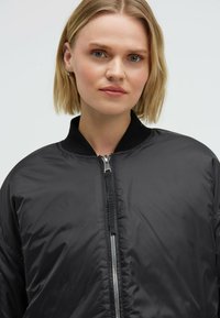 Schwarze Bomberjacke mit einem Reißverschluss vorne, geripptem Kragen und glatter Textur. Mit metallischem Reißverschluss und minimalistischen Design.