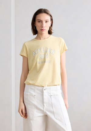 Νεαρή γυναίκα που φοράει κίτρινη ριγέ μπλούζα "Atelier 2010 Mos Mosh" και λευκό παντελόνι, στέκεται μπροστά σε απλό ανοιχτό φόντο.