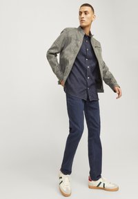 Gray suede bomber jacket, navy button-up shirt, dark jeans, and white sneakers with green and red accents. Model stands in a dynamic pose.