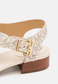 Sandal con tacco alto beige con tomaia in tessuto testurizzato caratterizzata da un motivo a logo marrone, fibbia color oro e tacco in legno.