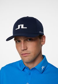Mörkblå keps med en vit logotyp på framsidan, strukturerad design och flexibel passform. Matchad med en ljusblå skjorta med krage.