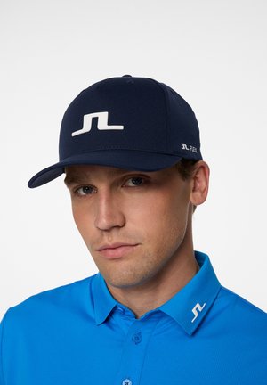 Navy blue cap with a white logo on the front, structured design, and a flexible fit. Paired with a light blue collared shirt.