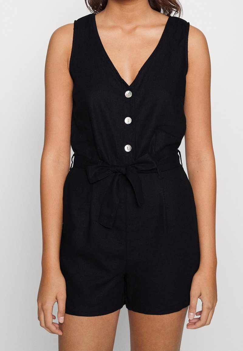 Combinaison en lin noir avec un décolleté en V profond, cinq boutons argentés sur le devant, taille à nouer ajustable et short. Design minimaliste, texture légère.