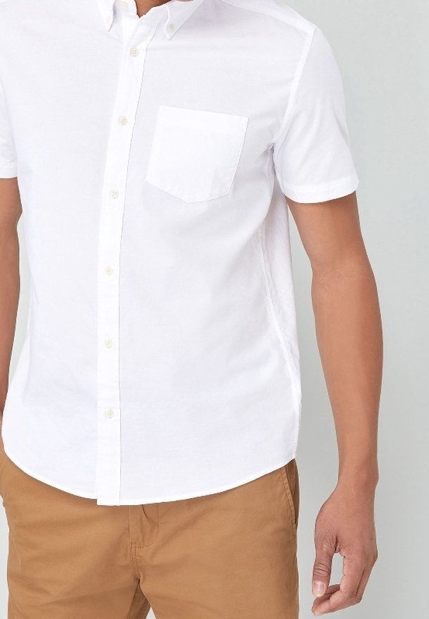 Homme portant une chemise blanche à manches courtes avec une poche poitrine et un pantalon beige, debout devant un fond uni.