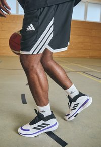 Melni basketbola šorti ar baltām sānu svītrām, apvienoti ar baltām zeķēm un melniem un violetiem basketbola apaviem, stāv uz sporta zāles grīdas.