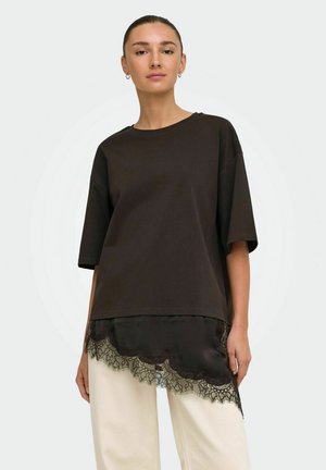 Mujer con una blusa negra de manga corta y suelta, superpuesta sobre un camisón de satén negro con encaje, combinada con pantalones de color crema.